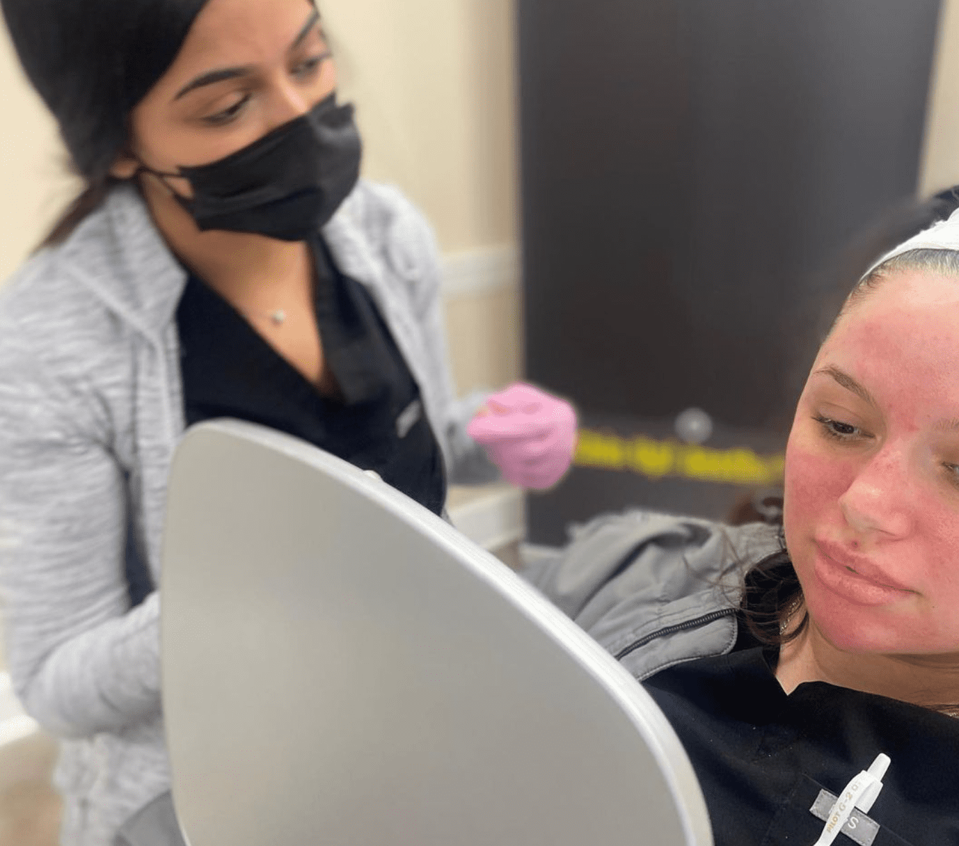 A woman with a headband and pink gloves performs an aesthetic skincare treatment on another woman who is lying down and looking at a mirror. The person performing the treatment, at what could be the best medspa in town, is wearing a black mask. Medspa near me