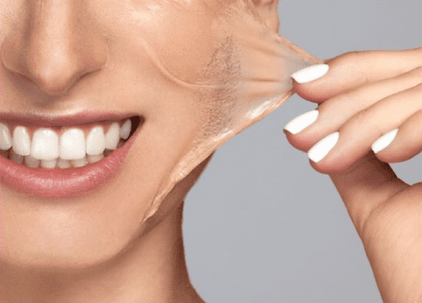 Close-up of a person with white teeth and white-painted nails peeling off a clear facial mask, revealing a glowing complexion. The lower half of their smiling face is visible, showing the results of their recent visit to a medspa. The background is gray. Medspa near me