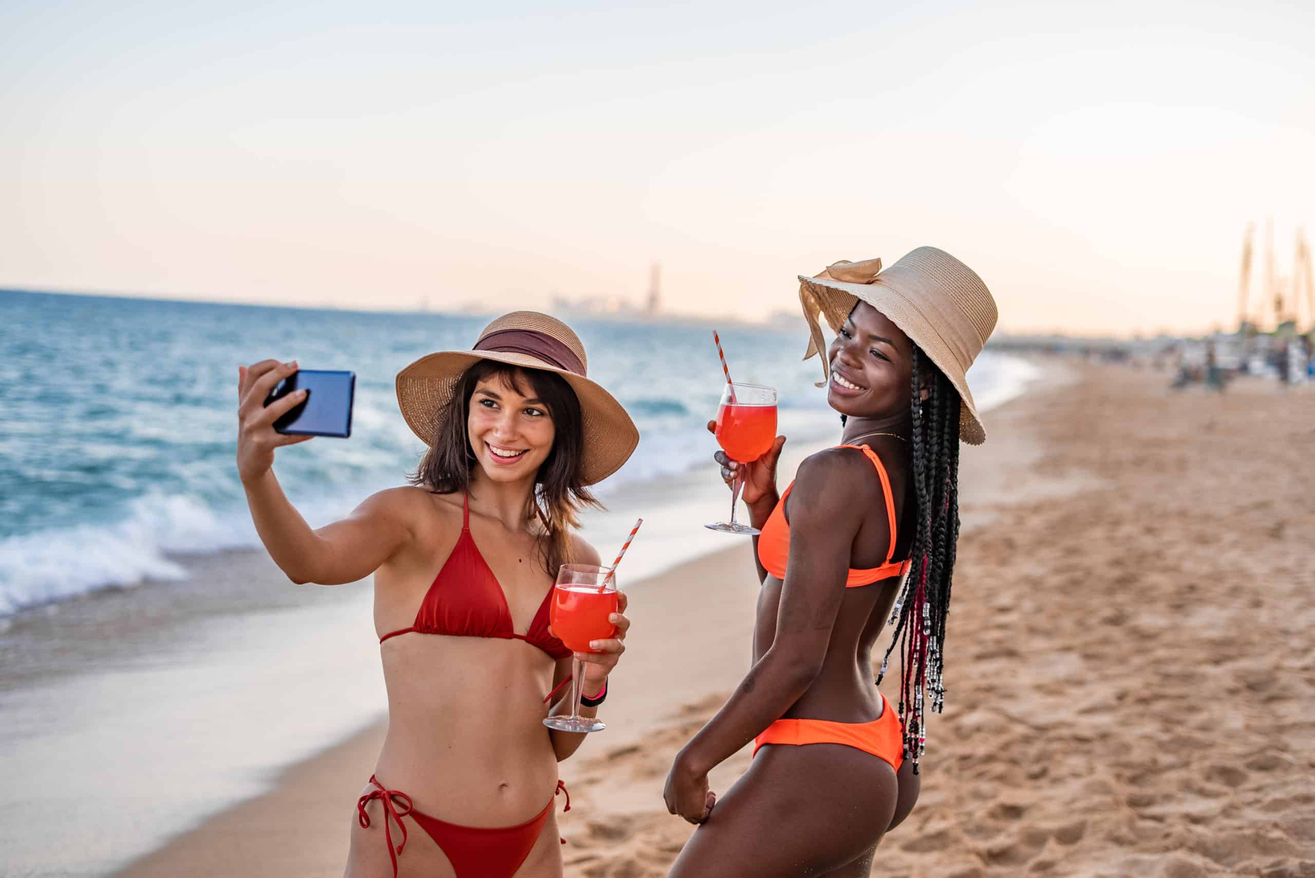 Two women in bikinis, wearing sun hats, smile as they take a selfie on a sandy beach near their favorite medspa. Both hold red cocktails with straws. The ocean waves gently lap the shore in the background, and the sky shows a soft, evening glow. Medspa near me