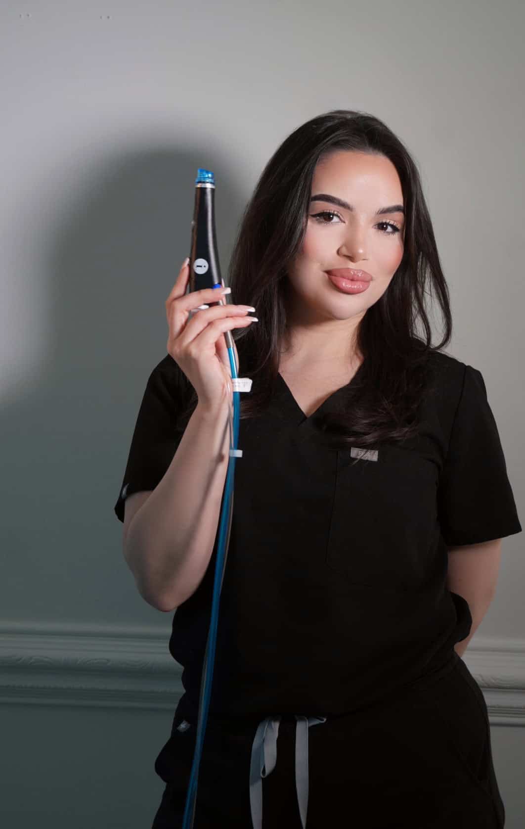 Woman holding a beauty device, wearing black scrubs.