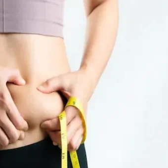 A person wearing a pink sports bra and black leggings pinches the skin on their waist with one hand while holding a yellow measuring tape around their abdomen with the other hand, perhaps considering a visit to the best medical spa near me. The background is plain and light-colored. Medspa near me