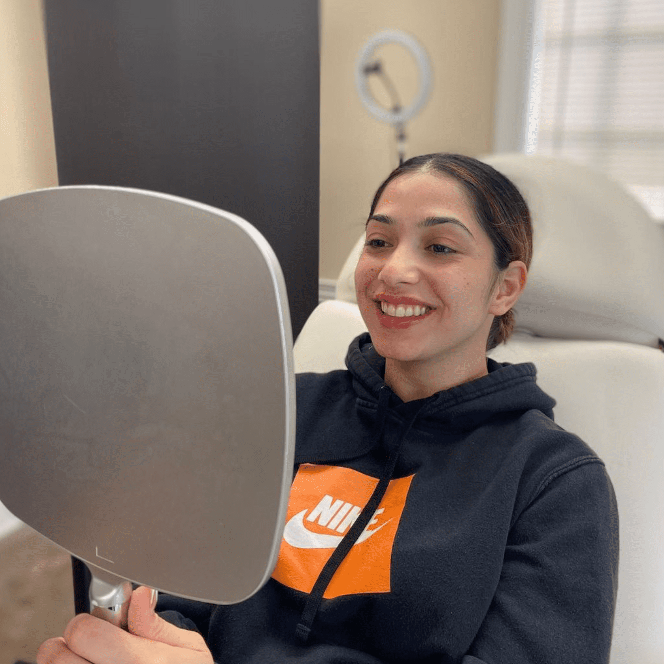 A woman is sitting in a chair and smiling at her reflection in a hand-held mirror. She is wearing a black hoodie with an orange and white Nike logo. Behind her, there’s a ring light and a window with blinds, capturing the serene ambiance often found in the best medical spas. Medspa near me