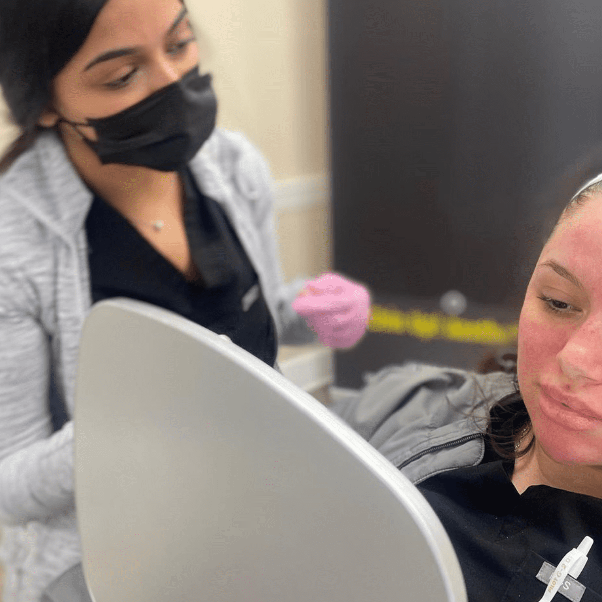 A woman with a headband and pink gloves performs an aesthetic skincare treatment on another woman who is lying down and looking at a mirror. The person performing the treatment, at what could be the best medspa in town, is wearing a black mask. Medspa near me