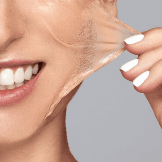 Close-up of a person with white teeth and white-painted nails peeling off a clear facial mask, revealing a glowing complexion. The lower half of their smiling face is visible, showing the results of their recent visit to a medspa. The background is gray. Medspa near me