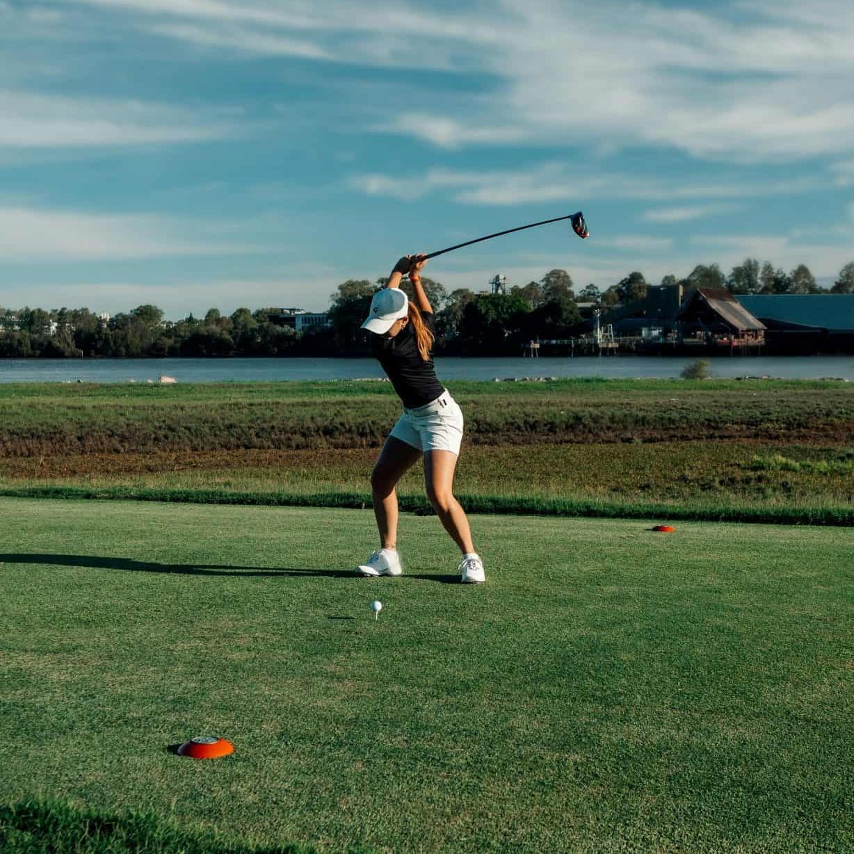 A golfer wearing a white cap, black shirt, and white shorts is in mid-swing on a green golf course under a partly cloudy sky. The serene background features a body of water and trees, reminiscent of the tranquility you'd find at a medspa near me. Medspa near me