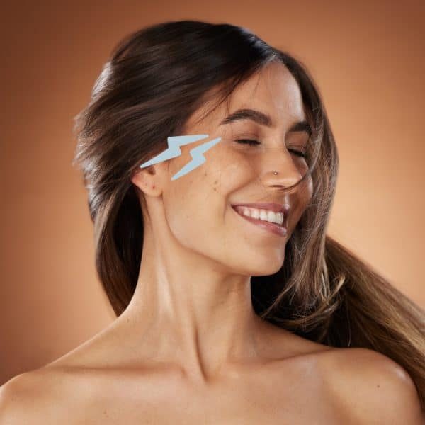 a woman with long brown hair smiles with eyes closed against a brown background. she has two blue lightning bolt stickers on her right cheek. she wears a nose ring and no visible clothing, giving an impression of natural beauty and joy. medspa near me botox on eyes