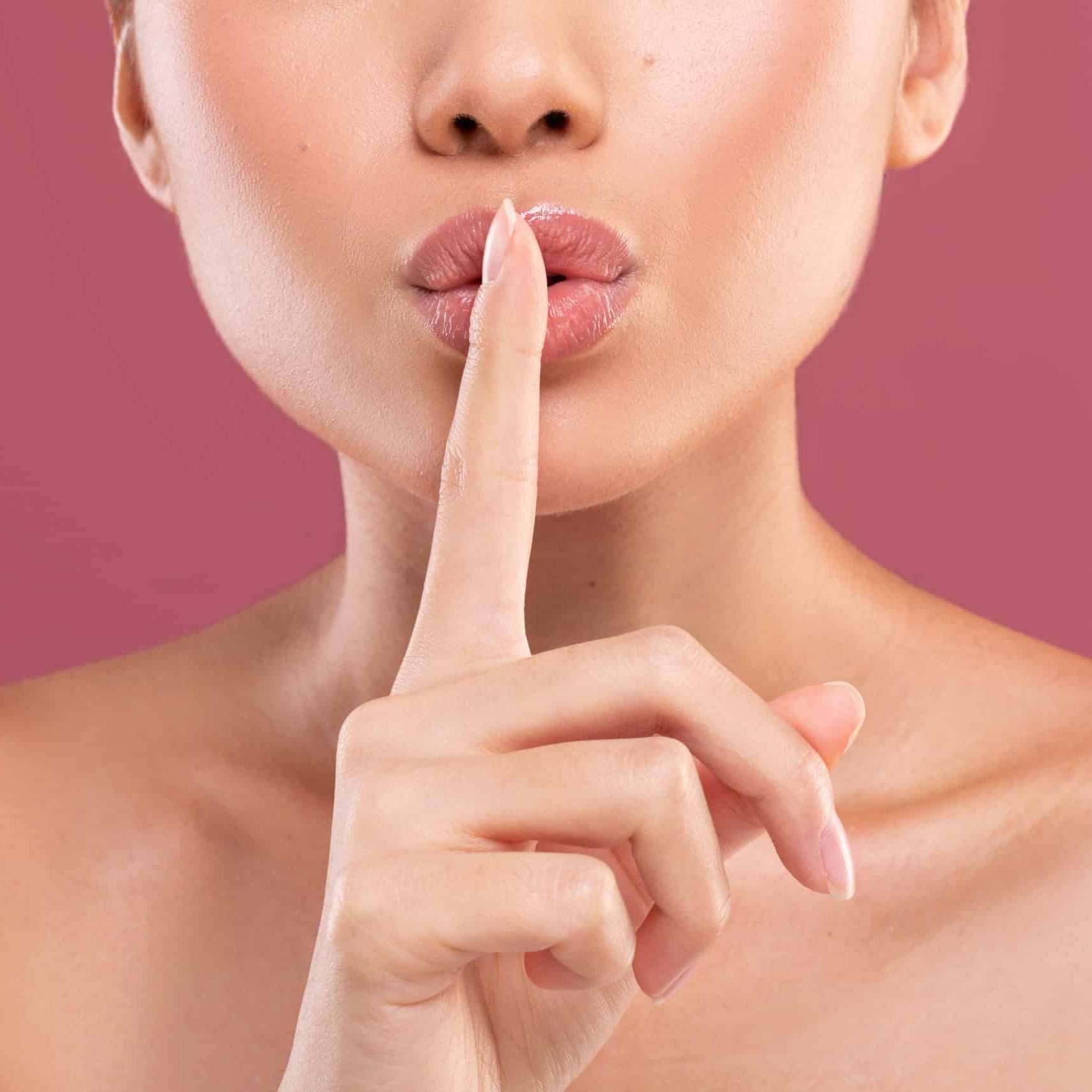 Close-up of a woman with a light skin tone pressing her index finger to her lips, which are slightly puckered, signaling for silence. The background is a solid pink color. Her shoulders are bare, evoking the soothing ambiance of a medspa. Medspa near me