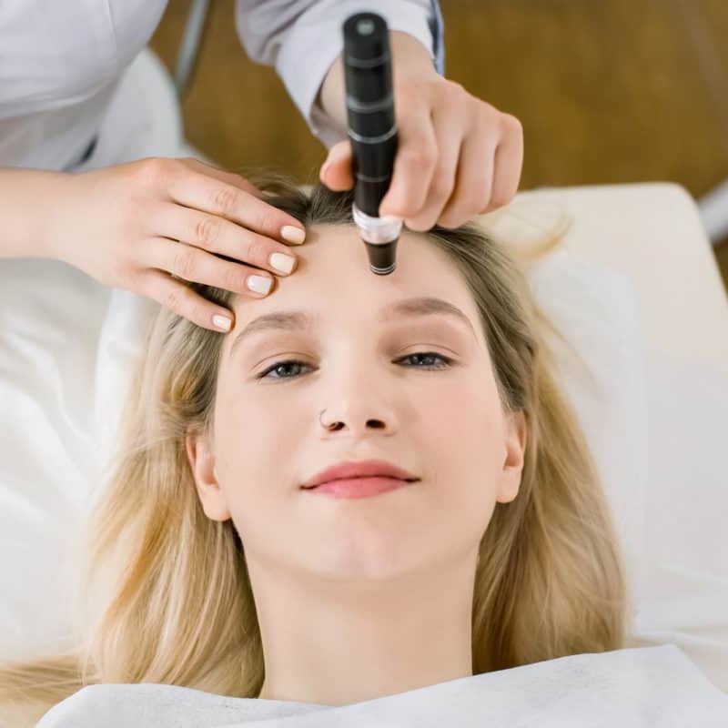 Dermatology and cosmetology clinic. Cropped top view of face of pretty blond woman during skin diagnostic. Hands of female doctor using a professional dermatoscope while doing the skin examination microneedling patient