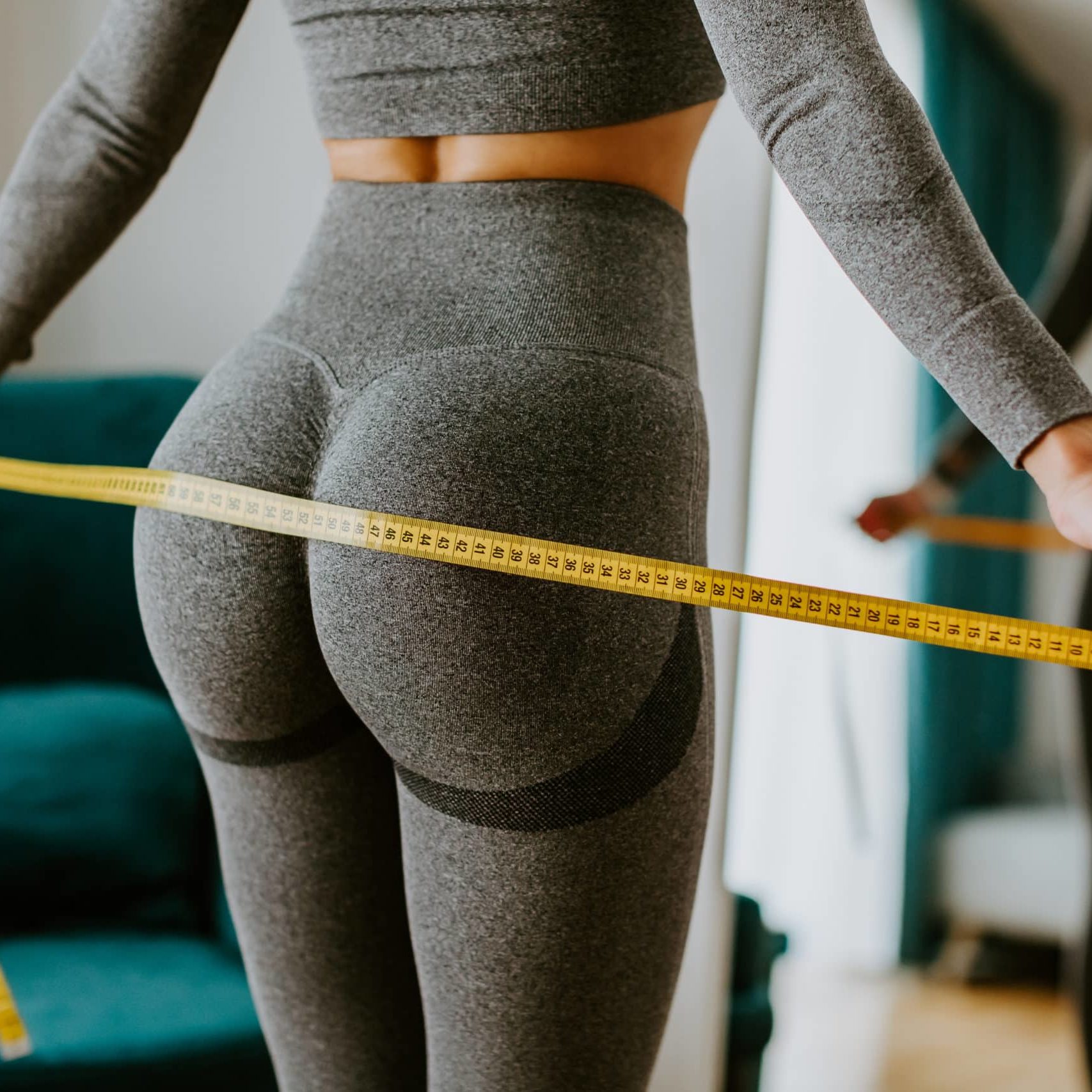 Fitness Woman Measuring body With Tape Standing In Front Of Mirror At Home. Beautiful woman checking success of weight loss program and measuring butt and hips in front of mirror in living room. Slimming Concept. Selective Focus.
