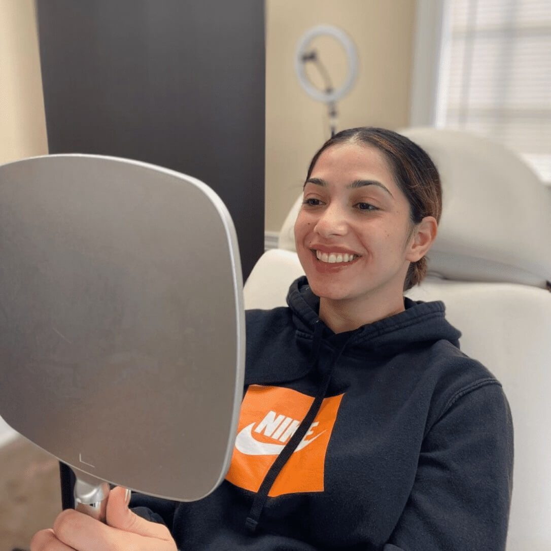 A woman is sitting in a chair and smiling at her reflection in a hand-held mirror. She is wearing a black hoodie with an orange and white Nike logo. Behind her, there’s a ring light and a window with blinds, capturing the serene ambiance often found in the best medical spas. Medspa near me A woman is sitting in a chair and smiling at her reflection in a hand-held mirror. She is wearing a black hoodie with an orange and white Nike logo. Behind her, there’s a ring light and a window with blinds, capturing the serene ambiance often found in the best medical spas. Medspa near me
