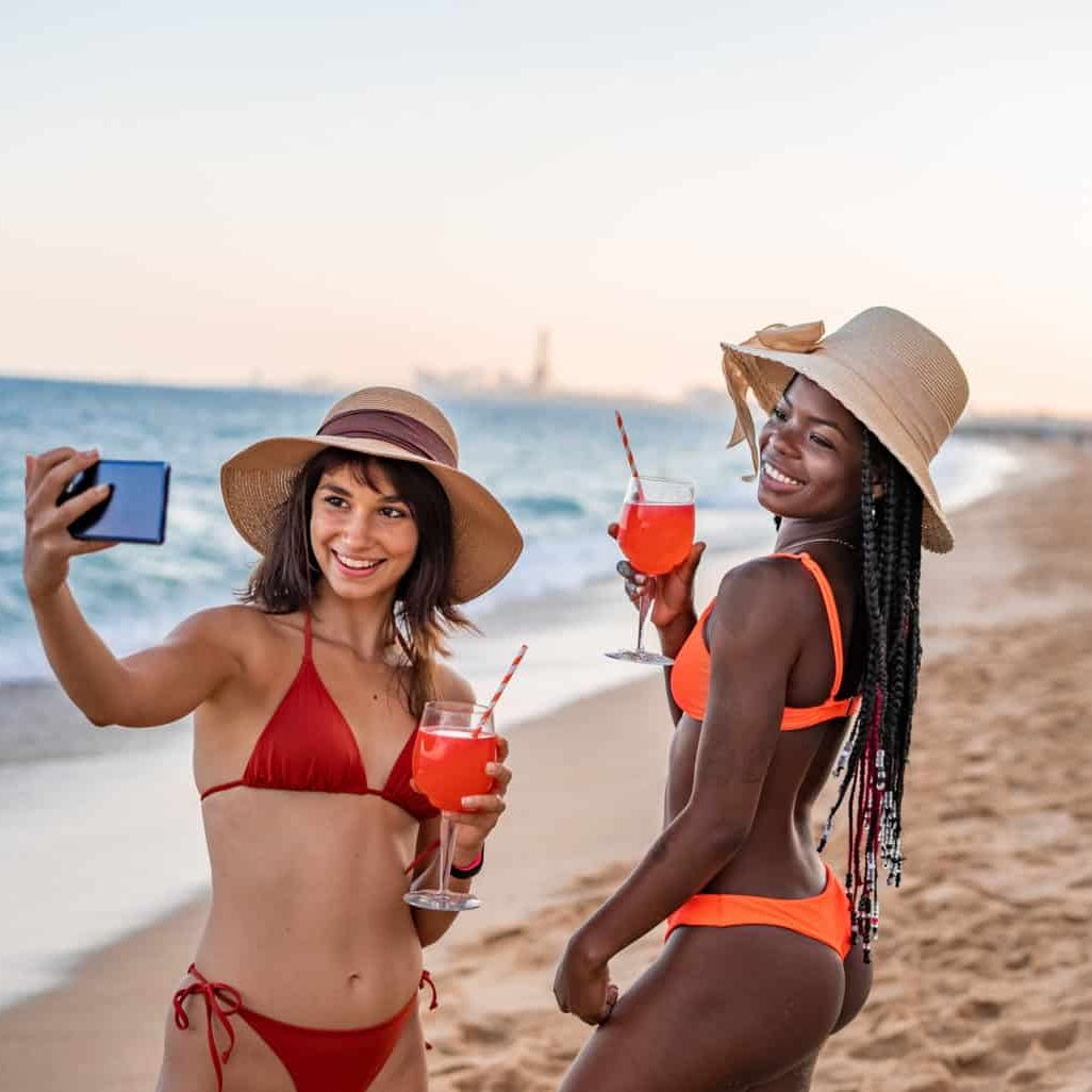 Two women in bikinis, wearing sun hats, smile as they take a selfie on a sandy beach near their favorite medspa. Both hold red cocktails with straws. The ocean waves gently lap the shore in the background, and the sky shows a soft, evening glow. Medspa near me