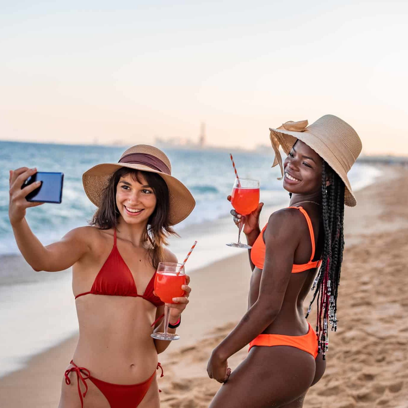 Charming young fit diverse women in swimwear and hats with drinks taking selfie on mobile phone while spending summer evening together on sandy beach near ocean