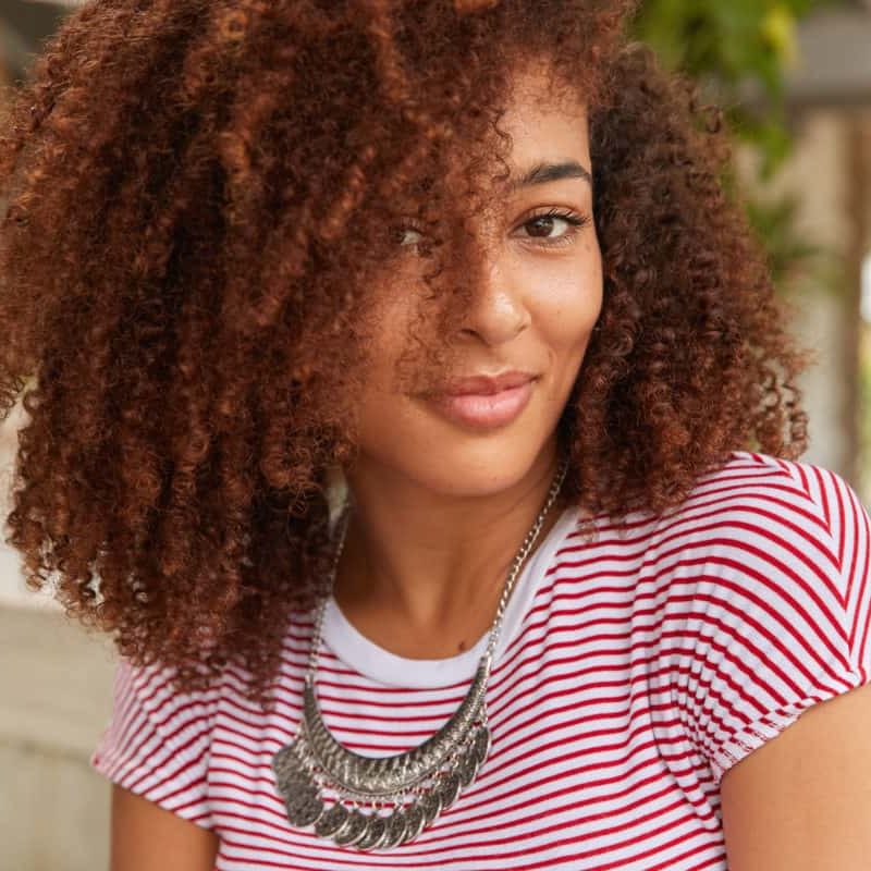 Headshot of attractive woman with curly hair, looks positively at camera, satisfied with something, wears striped t shirt and silver jewellery, models in restaurant. Close up of dark skinned girl