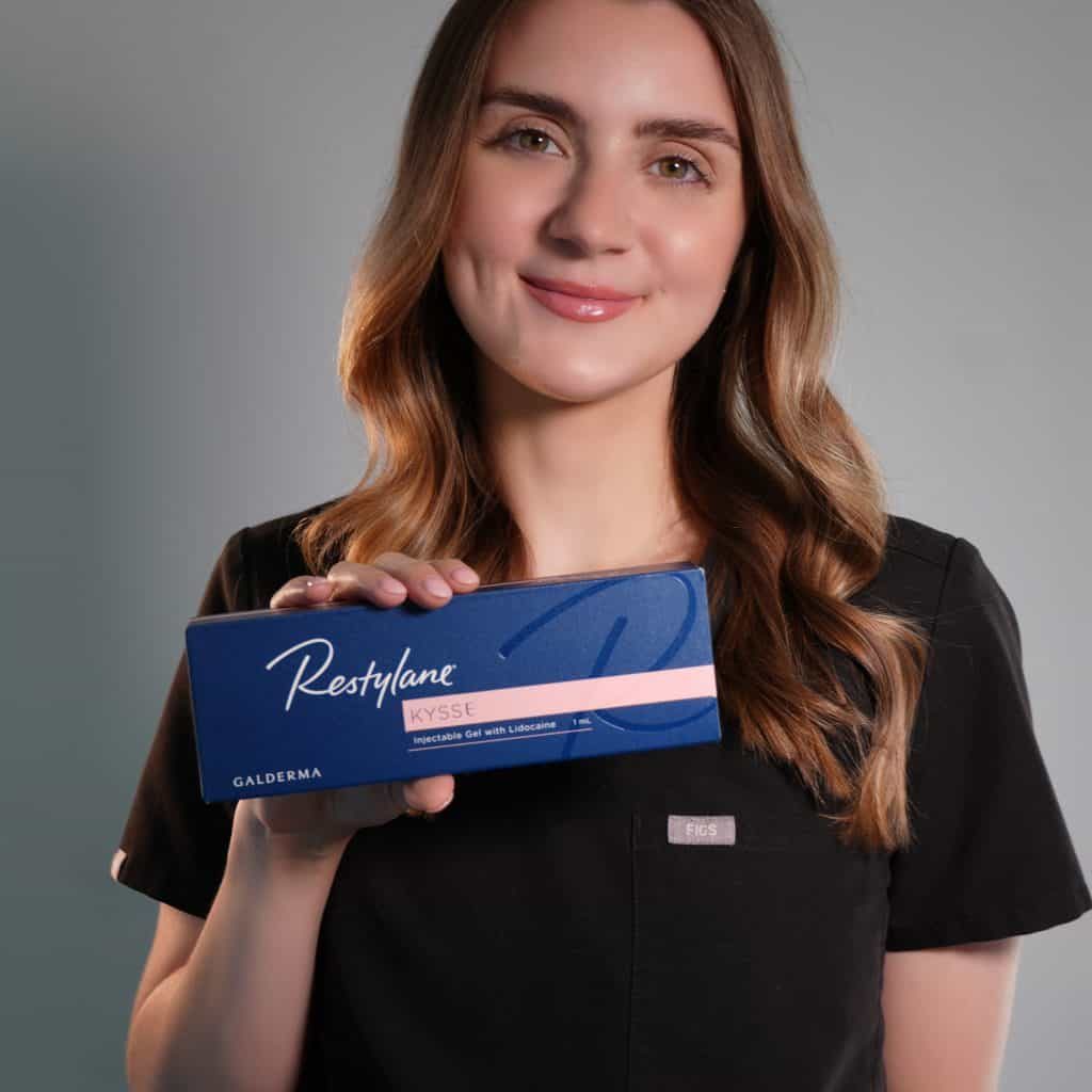 a woman with long brown hair, wearing black scrubs, smiles and holds a blue box labeled "restylane kysse" in front of a neutral background. medspa near me