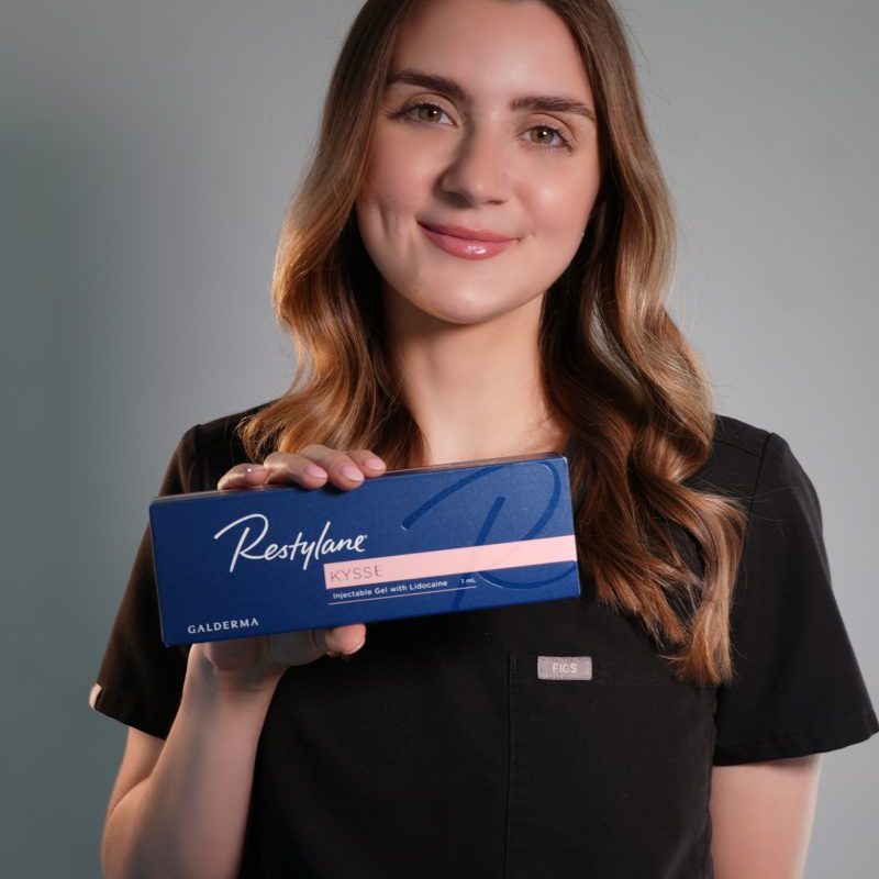a woman with long brown hair, wearing black scrubs, smiles and holds a blue box labeled "restylane kysse" in front of a neutral background. medspa near me