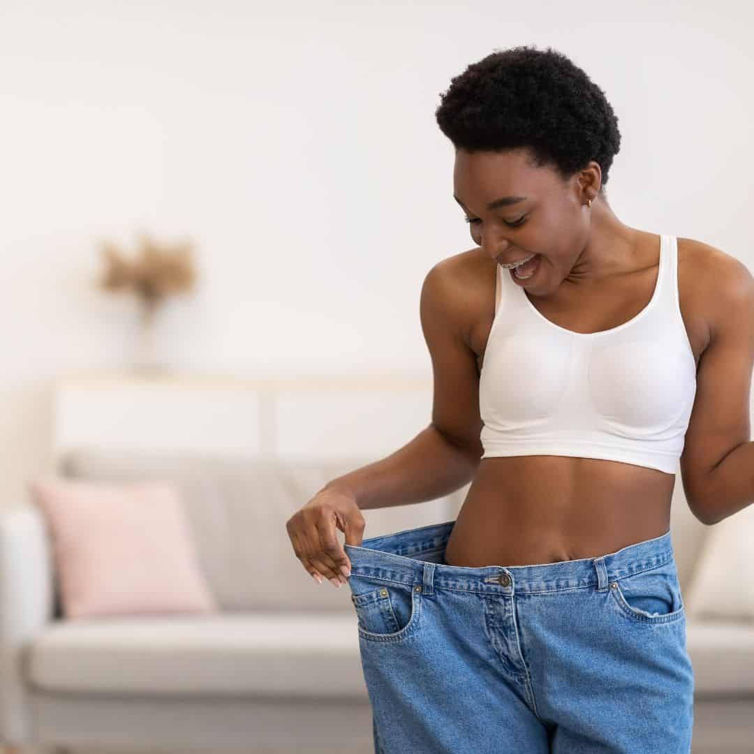 A person with short curly hair and wearing a white sports bra is looking down and smiling, holding out the waistband of their oversized jeans proudly. Indoors, with a cozy couch and pillows in the background, they radiate confidence reminiscent of someone fresh from a medspa visit. Medspa near me